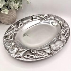 May include: Oval silver-colored serving tray with a detailed border of seashells. The tray has a smooth, reflective surface and a raised edge. The decorative border features various types of seashells, creating a coastal or nautical theme.
