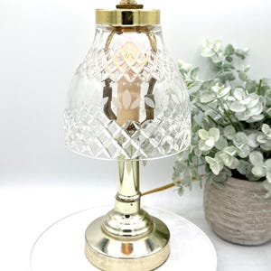 May include: A vintage-style table lamp with a clear, diamond-cut glass shade and a gold-colored metal base. The lamp features a decorative finial and a warm, soft glow. The lamp is approximately 10 inches tall.