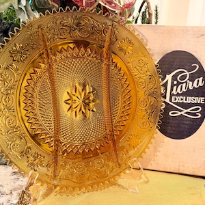 May include: A vintage amber-colored Tiara Exclusive divided serving dish. The dish features intricate embossed floral and geometric patterns, a starburst center, and a scalloped edge. The dish is displayed on a clear stand, with the original box visible.