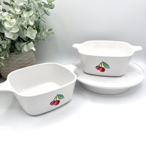 May include: Two white rectangular baking dishes with handles, each adorned with a cherry design. One dish rests on a white plate, while the other sits on a white surface. A decorative plant is in the background.