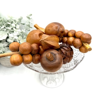 May include: A collection of wooden decorative objects in various shapes and sizes, including spheres, carved pieces, and a pinecone, displayed on a clear glass pedestal dish. The wooden pieces are in shades of brown and tan.