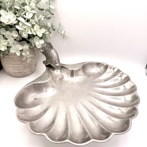 May include: A silver-colored, scallop-shaped serving dish with a fish-shaped handle. The dish has a fluted design and is made of metal. The decorative dish is suitable for serving appetizers or displaying small items. The dish is approximately 12 inches in diameter.