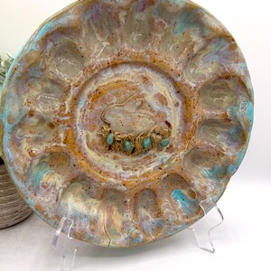 May include: A round ceramic serving platter with twelve circular indentations around the perimeter. The glaze features a marbled blend of tan, cream, and turquoise blue. The center displays a sculpted rabbit figure with a small nest of eggs.