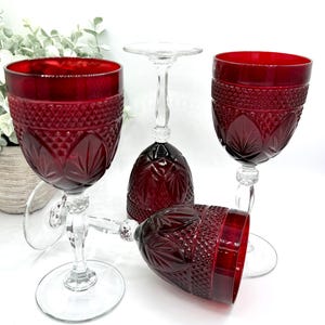 May include: A set of four deep red glass goblets featuring a textured diamond pattern on the upper bowl and a starburst design on the lower section. Each goblet has a clear glass stem and a circular base, showcasing a pressed glass construction.