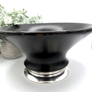 May include: A black decorative bowl with a wide, flared rim and a silver-colored base. The bowl has a smooth, matte finish and a slightly distressed appearance around the rim. The base is a shiny, metallic silver.