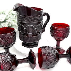 May include: A ruby red glass pitcher and matching goblets. The pitcher has a handle and a textured pattern. The goblets are on stems and have a similar design. The glassware is arranged on a white surface.