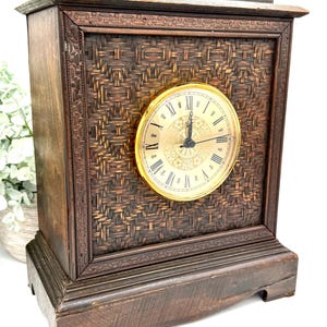 May include: A vintage wooden clock with a woven front panel and a gold-toned clock face. The clock has Roman numerals and ornate detailing, showcasing a classic design. The clock is a dark brown color.