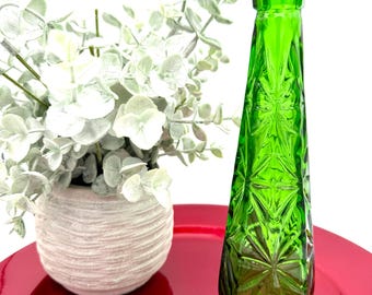 May include: A vibrant green glass vase with a starburst pattern, standing on a red surface. Beside it, a small white pot holds faux eucalyptus branches. The vase is approximately 10 inches tall.
