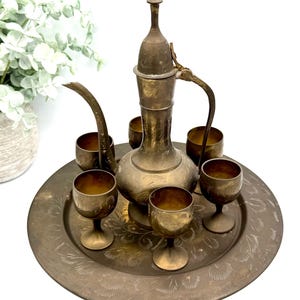 May include: Antique brass tea set with a decorative etched design. The set includes a tall, ornate teapot with a spout and handle, a round tray, and five small goblets. The set is displayed on a white surface.