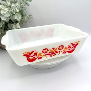May include: White rectangular baking dish with a red and orange floral and bird design. The dish has a handle on each end and is sitting on a white plate. The design features stylized birds and flowers.