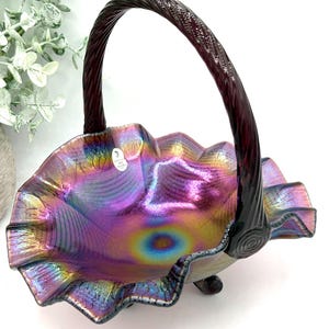 May include: A decorative glass basket featuring an iridescent finish with purple, blue, and gold hues. The bowl has a ruffled, crimped edge and a dark, twisted handle. A small white sticker with text is visible on the interior surface of the bowl.