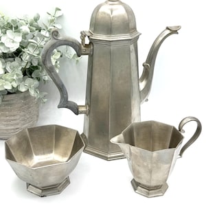 May include: A vintage silver tea set, including a tall coffee pot with a domed lid, a sugar bowl, and a creamer. The set has a geometric design and a curved handle on the coffee pot. The pieces are arranged on a white background.