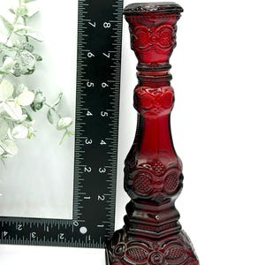 May include: A deep red glass candlestick holder with an intricate embossed pattern, standing approximately 8.5 inches tall as indicated by the black ruler placed alongside it. The piece features a square base and a cylindrical stem with decorative details.