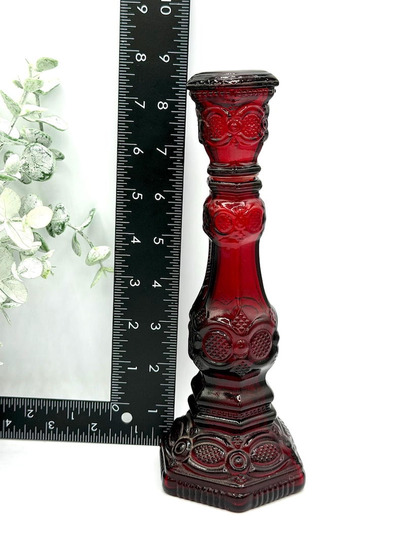 May include: A deep red glass candlestick holder with an intricate embossed pattern, standing approximately 8.5 inches tall as indicated by the black ruler placed alongside it. The piece features a square base and a cylindrical stem with decorative details.