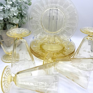 May include: A collection of vintage yellow glassware and plates. The set includes plates with a radial design, stemmed glasses, and footed glasses, all with etched floral patterns. The glassware is arranged on a white surface.