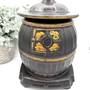 May include: A black ceramic container with a lid, featuring a decorative design with gold accents. The container has a cylindrical shape with a base and a lid. The design includes floral patterns in shades of yellow and orange.