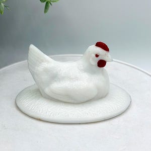 May include: A white ceramic hen figurine with a red comb and wattle. The hen is sitting on a white, round base with a textured pattern. The figurine is a decorative item, possibly for a kitchen or farmhouse decor.
