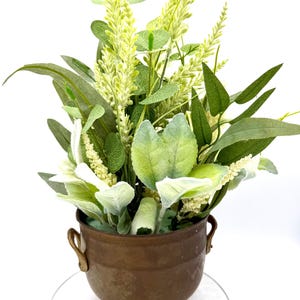 May include: A decorative arrangement of artificial greenery and flowers in a small, aged bronze-colored pot. The arrangement features various shades of green leaves and light green, textured floral accents. The pot has two handles and rests on three ornate feet.