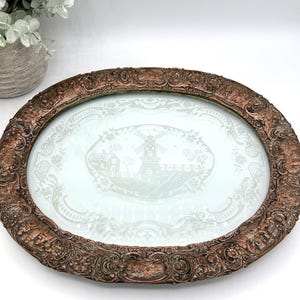 May include: Oval antique serving tray with a detailed bronze-colored frame. The tray's center features a light blue glass insert with a white windmill and landscape design. The frame has ornate scrollwork and relief details.
