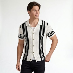 Op de afbeelding: Een crème-kleurig overhemd met korte mouwen, zwarte verticale strepen en een kraag. Het overhemd heeft horizontale zwarte strepen op de mouwen. Het model draagt een wit ondershirt en een donkerblauwe jeans. Het overhemd is dichtgeknoopt.