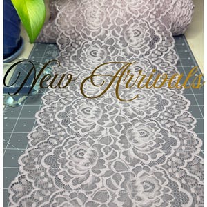 Può includere: Un rotolo di tessuto in pizzo floreale grigio chiaro. Il pizzo presenta un motivo ripetuto di rose e altri elementi floreali. Le parole "New Arrivals" sono scritte in oro sull'immagine. Il pizzo è su un tappetino da taglio grigio.