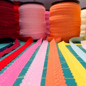 May include: A collection of colorful ric rac trim in various shades, including red, pink, orange, yellow, and blue. The trim is displayed on spools and laid out flat, showcasing the scalloped edges. The background is a green cutting mat.