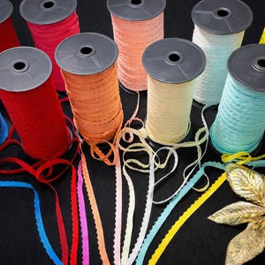 Op de afbeelding: Een verzameling kleurrijke lintspoelen in verschillende tinten, waaronder blauw, rood, oranje en geel. De spoelen zijn gerangschikt op een zwart oppervlak, met het lint afgewikkeld, waarbij de getextureerde randen zichtbaar zijn. Er is ook een gouden decoratieve bloem aanwezig.