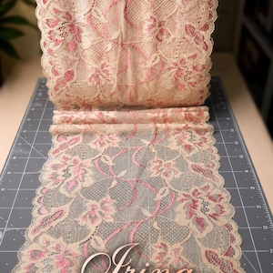 May include: A roll of beige lace fabric featuring a floral pattern with pink accents. The lace has a scalloped edge and a delicate mesh texture. The word "Irina" is printed in a cursive font at the bottom of the image over the fabric.