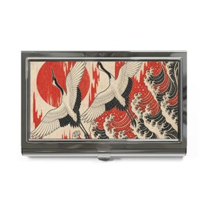 May include: A rectangular metal business card case featuring a graphic print of two white cranes with black wing tips flying over stylized black waves against a textured cream background with a large red sun and red vertical streaks.