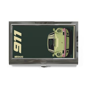 May include: A rectangular metal business card holder featuring a dark green background with a light green sports car illustration. The text 911 is printed in large cream letters, and the word SINGER is printed in smaller cream letters.