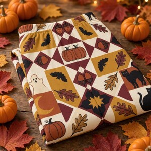 Peut inclure: Pantalon de détente plié avec un motif géométrique en losanges crème, jaune moutarde et bordeaux foncé. L'imprimé présente des illustrations de citrouilles orange, chauves-souris noires, chats noirs, glands bruns, feuilles de chêne et un fantôme blanc.