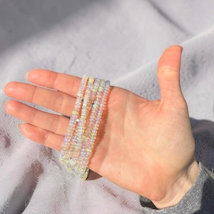May include: Several beaded bracelets with iridescent, opal-like beads are displayed. The bracelets are held in a hand against a soft, gray background. The beads appear to be small and round, with a subtle shimmer.