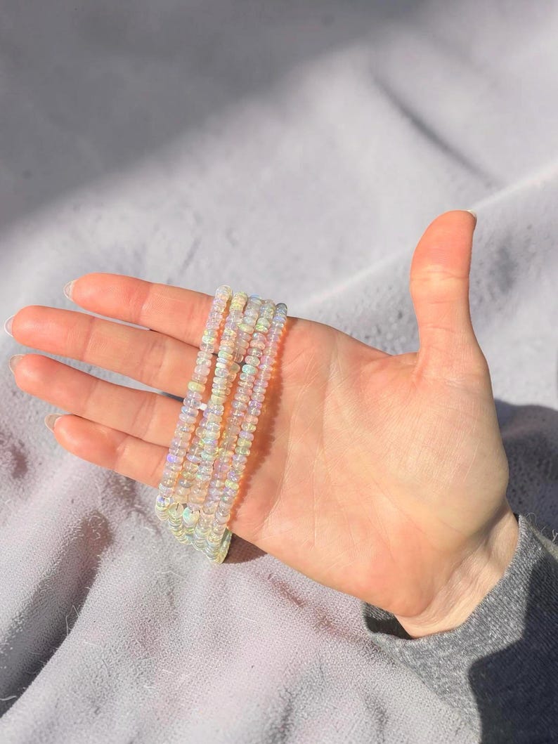 May include: Several beaded bracelets with iridescent, opal-like beads are displayed. The bracelets are held in a hand against a soft, gray background. The beads appear to be small and round, with a subtle shimmer.