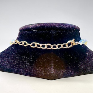 May include: A gold-colored chain necklace with iridescent beads. The necklace is displayed on a black velvet jewelry stand. The clasp and chain links are visible, showcasing the necklace's design and construction.