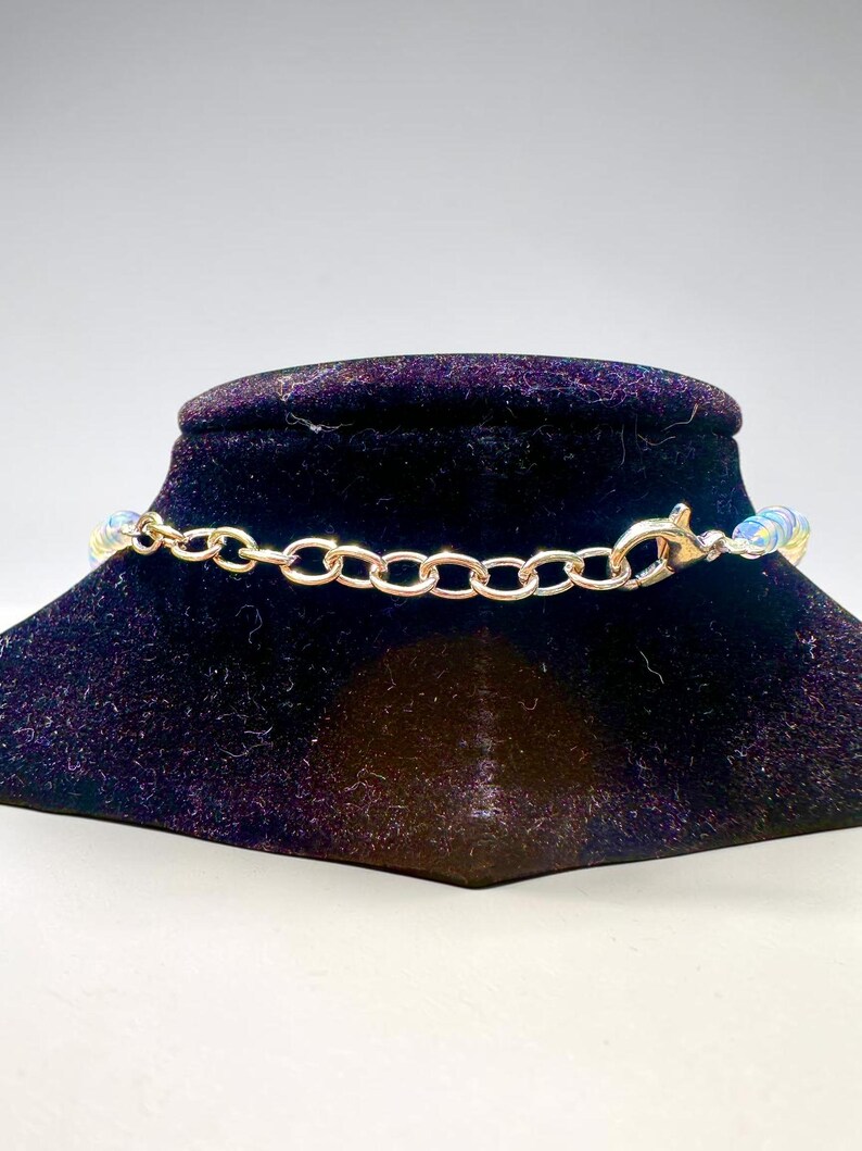 May include: A gold-colored chain necklace with iridescent beads. The necklace is displayed on a black velvet jewelry stand. The clasp and chain links are visible, showcasing the necklace's design and construction.