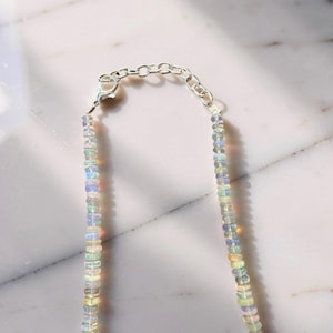 May include: A delicate necklace featuring small, iridescent opal beads strung together. The necklace has a silver-toned chain and clasp. The opal beads display a range of colors, including blue, green, and yellow, creating a subtle, shimmering effect.