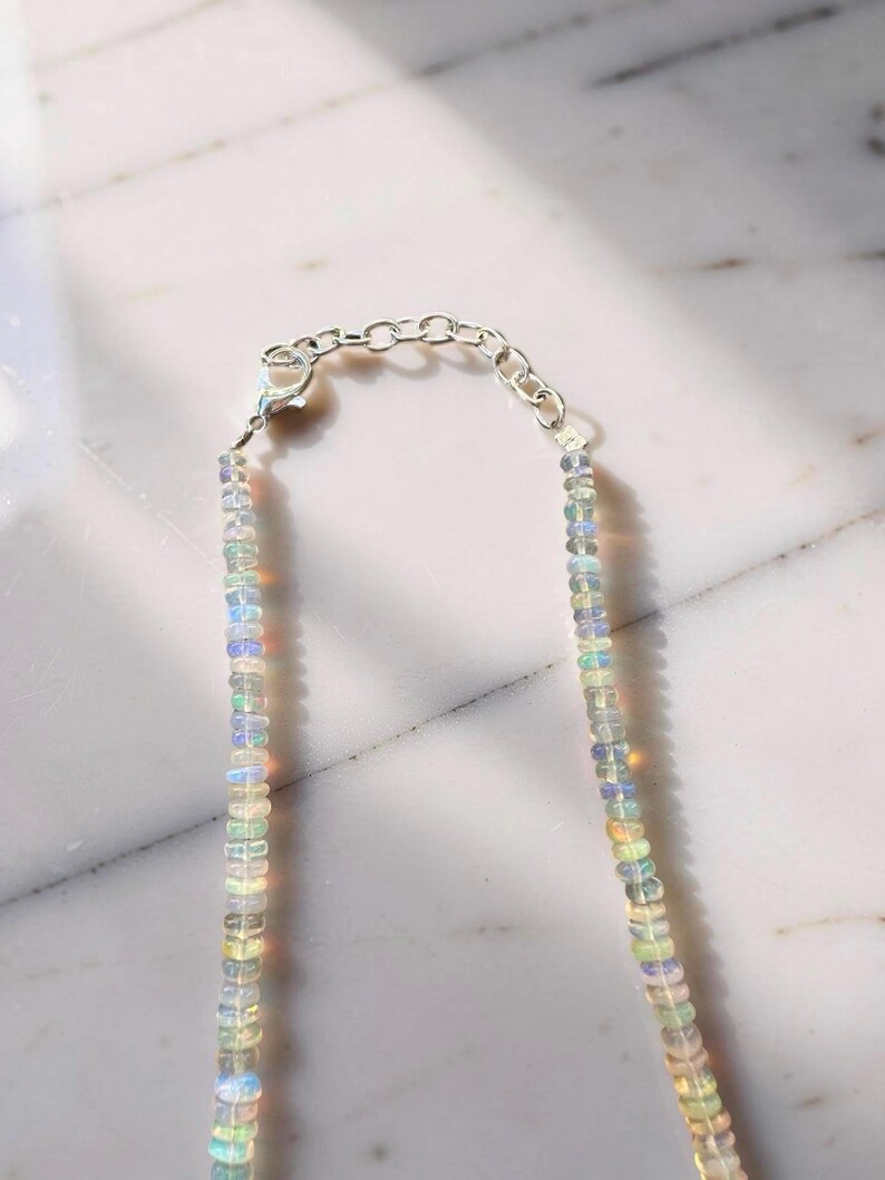 May include: A delicate necklace featuring small, iridescent opal beads strung together. The necklace has a silver-toned chain and clasp. The opal beads display a range of colors, including blue, green, and yellow, creating a subtle, shimmering effect.