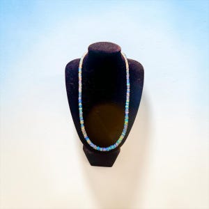 May include: A delicate necklace composed of small, iridescent beads, displayed on a black velvet jewelry bust. The beads exhibit a range of colors, including blue, green, and yellow, creating a shimmering effect. The necklace is a jewelry item.