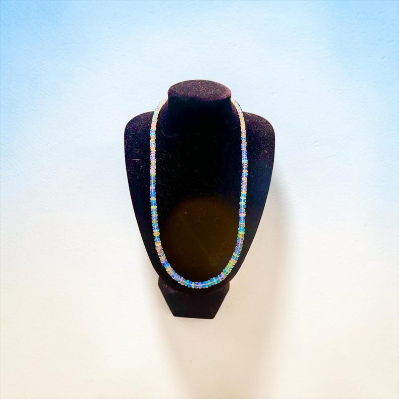 May include: A delicate necklace composed of small, iridescent beads, displayed on a black velvet jewelry bust. The beads exhibit a range of colors, including blue, green, and yellow, creating a shimmering effect. The necklace is a jewelry item.