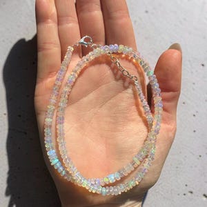 May include: A delicate necklace composed of small, iridescent opal beads. The necklace features a silver clasp and chain, and the beads display a range of colors, including white, blue, and green. The jewelry is displayed on a light background.