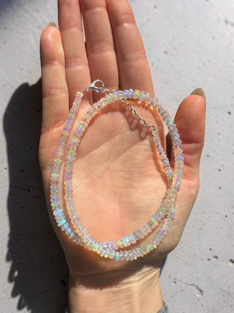 May include: A delicate necklace composed of small, iridescent opal beads. The necklace features a silver clasp and chain, and the beads display a range of colors, including white, blue, and green. The jewelry is displayed on a light background.
