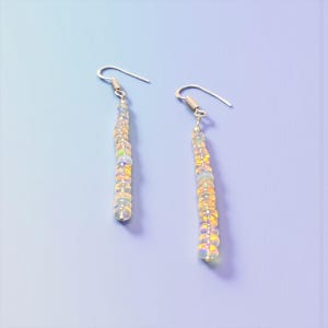 May include: A pair of dangle earrings with silver hooks. Each earring features a strand of iridescent, round beads in shades of yellow, white, and opal. The earrings are displayed against a light blue background.
