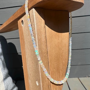 May include: A delicate opal bead necklace. The necklace features small, round, iridescent opal beads strung together. The beads display a range of colors, including white, blue, green, and yellow. The necklace is displayed against a wooden chair.