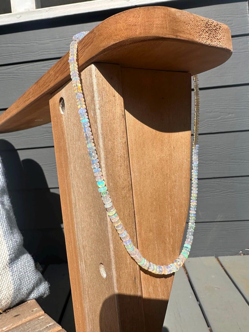 May include: A delicate opal bead necklace. The necklace features small, round, iridescent opal beads strung together. The beads display a range of colors, including white, blue, green, and yellow. The necklace is displayed against a wooden chair.
