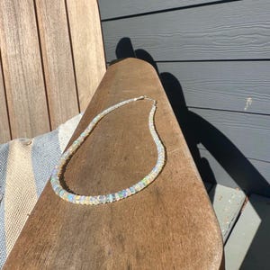 May include: A delicate opal necklace with iridescent beads. The necklace is resting on a wooden chair. The beads have a range of colors, including white, blue, and green. The necklace is a beautiful piece of jewelry.