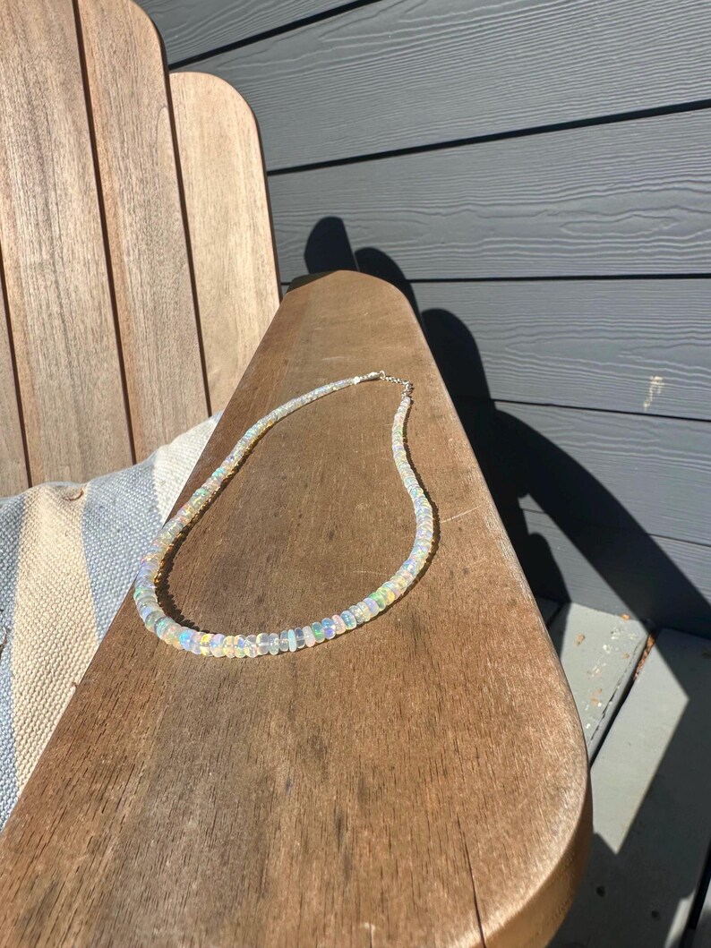 May include: A delicate opal necklace with iridescent beads. The necklace is resting on a wooden chair. The beads have a range of colors, including white, blue, and green. The necklace is a beautiful piece of jewelry.