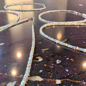 May include: Several opal necklaces with iridescent colors are displayed on a dark, speckled surface. The necklaces are composed of small, round opal beads strung together, with a silver clasp. The necklaces are in various positions, showcasing their design.