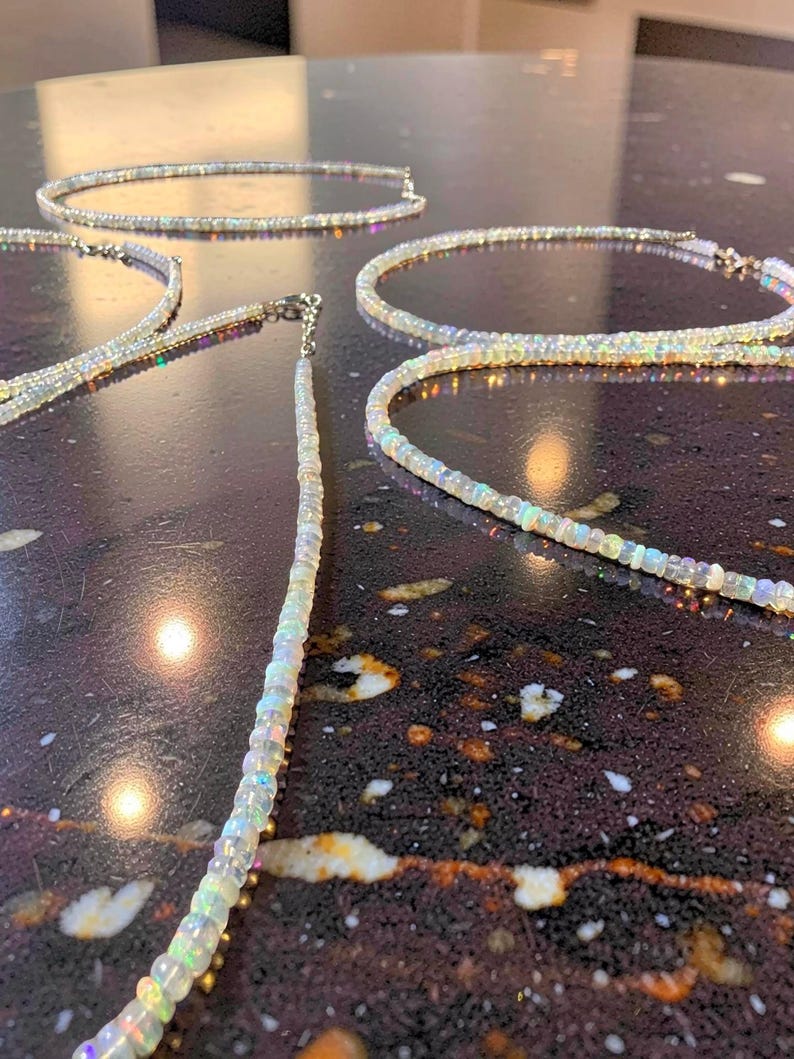 May include: Several opal necklaces with iridescent colors are displayed on a dark, speckled surface. The necklaces are composed of small, round opal beads strung together, with a silver clasp. The necklaces are in various positions, showcasing their design.