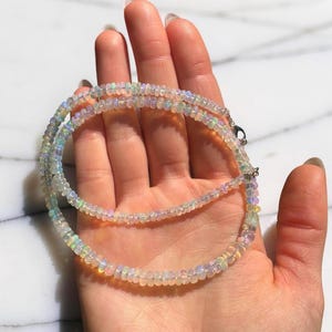 May include: A delicate necklace composed of small, iridescent opal beads. The necklace is displayed on a hand, showcasing the subtle play of colors within the opals. The beads are strung together, creating a layered effect. The necklace is a piece of jewelry.