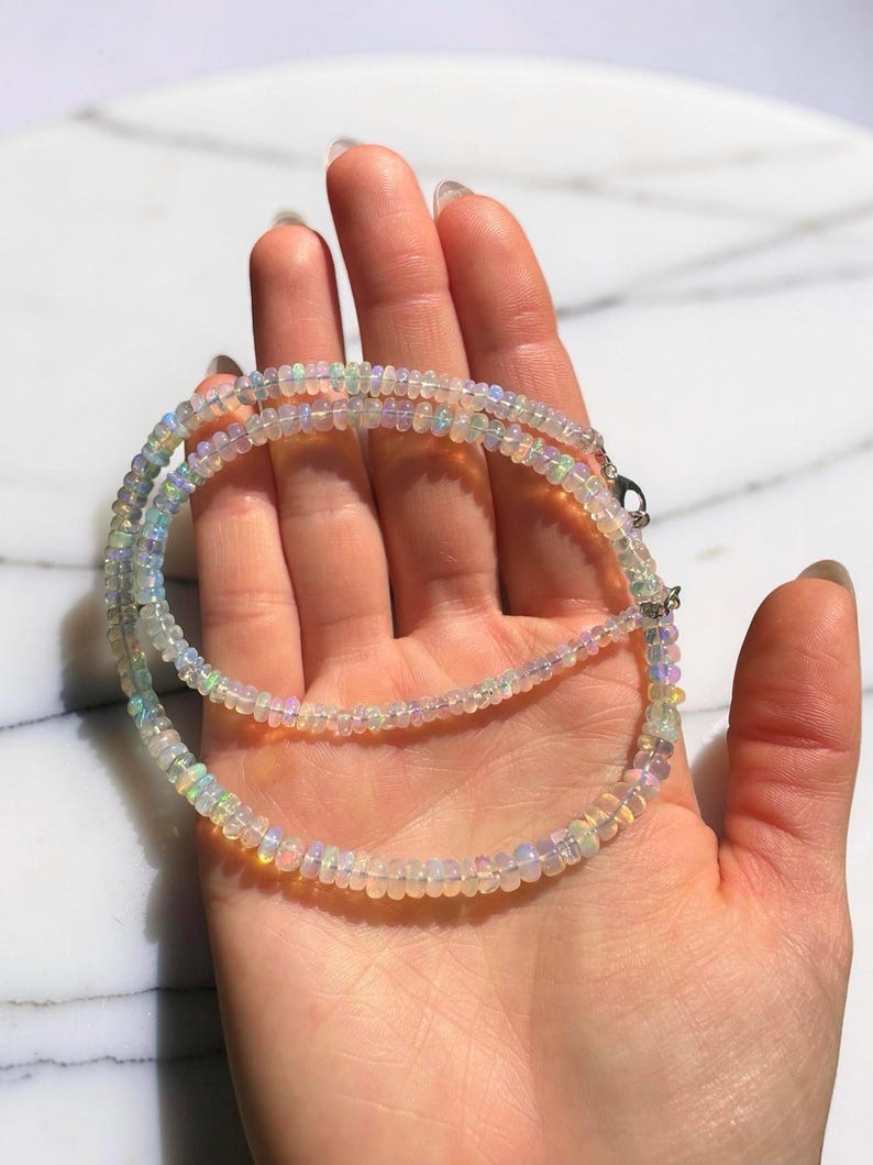 May include: A delicate necklace composed of small, iridescent opal beads. The necklace is displayed on a hand, showcasing the subtle play of colors within the opals. The beads are strung together, creating a layered effect. The necklace is a piece of jewelry.